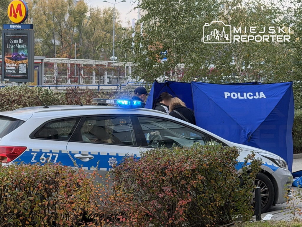 Policyjny radiowóz i niebieski parawan z napisem „POLICJA” przy stacji metra Ursynów, wśród jesiennych drzew.
