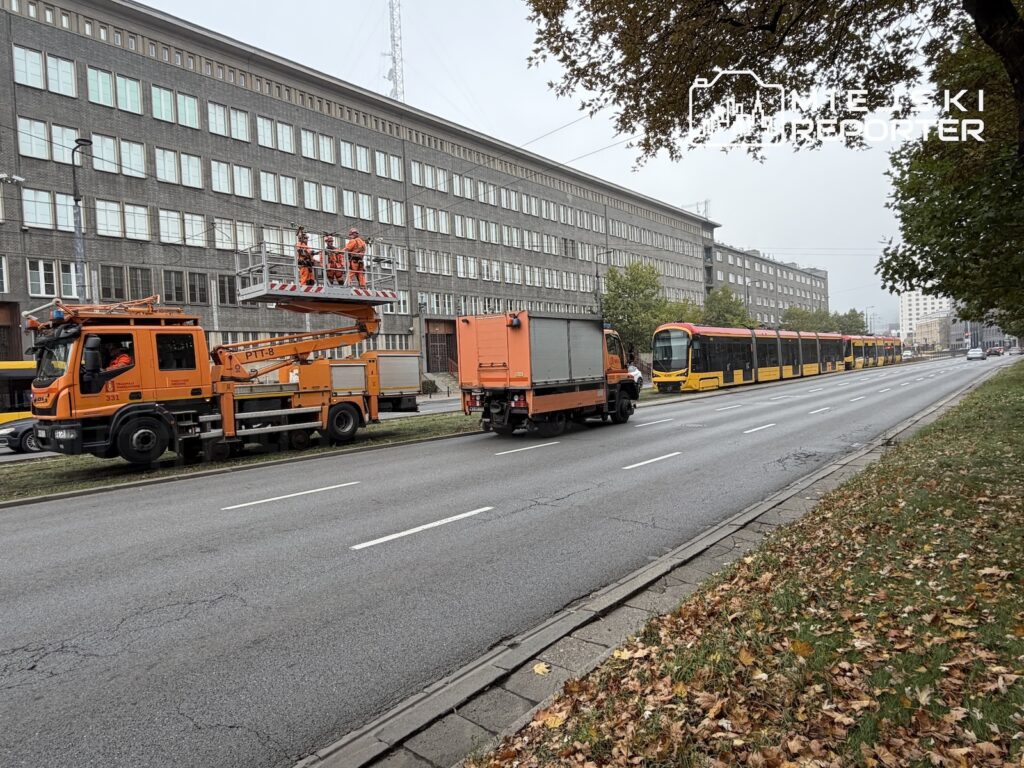 Pracownicy na podnośniku naprawiają sieć trakcyjną obok pomarańczowych samochodów technicznych i stojącego tramwaju.