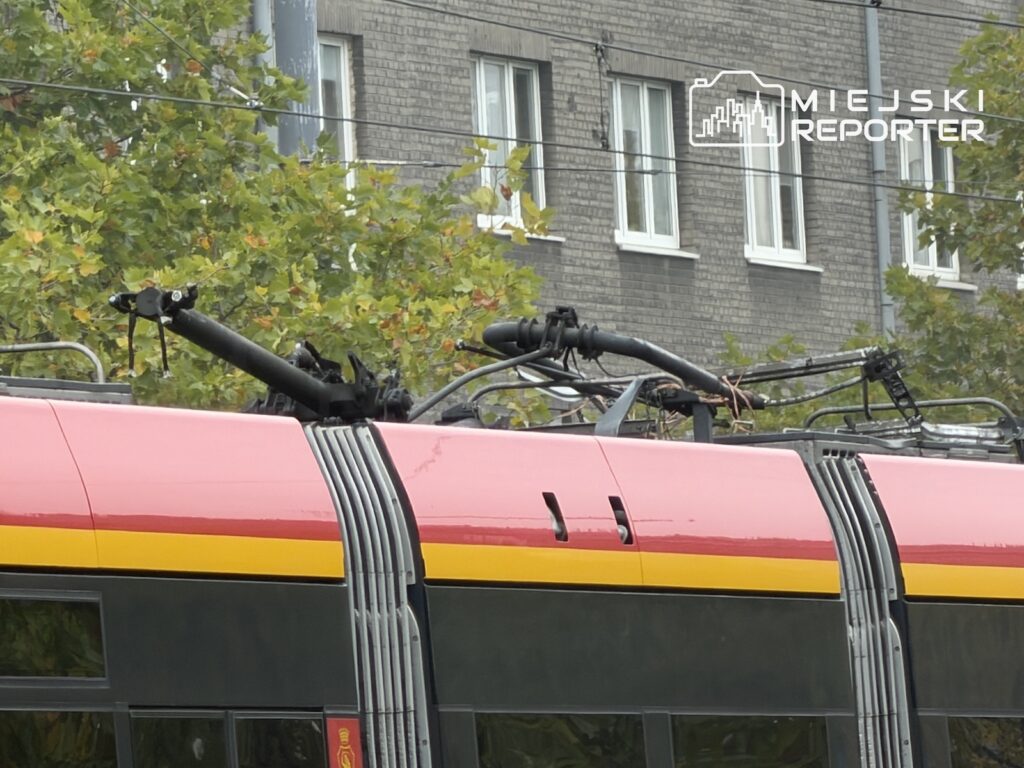 Zbliżenie na dach różowo-żółtego tramwaju z widocznym pantografem, na tle drzew i kamienicy.