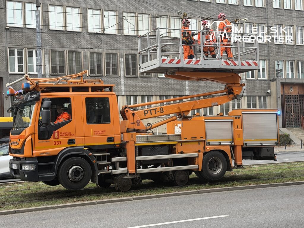 Pracownicy w odblaskowych kombinezonach naprawiają sieć trakcyjną z podnośnika przy pomarańczowym samochodzie serwisowym.