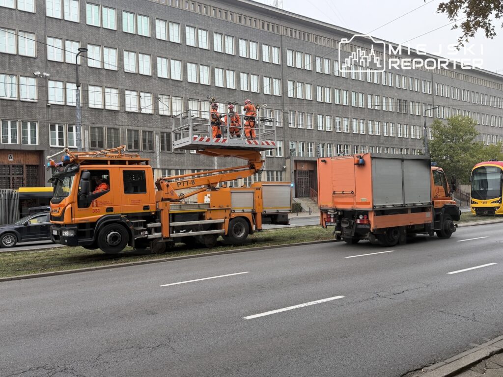 Pracownicy w pomarańczowych kombinezonach naprawiają sieć trakcyjną z podnośnika przy ulicy, obok stoi tramwaj i ciężarówki.