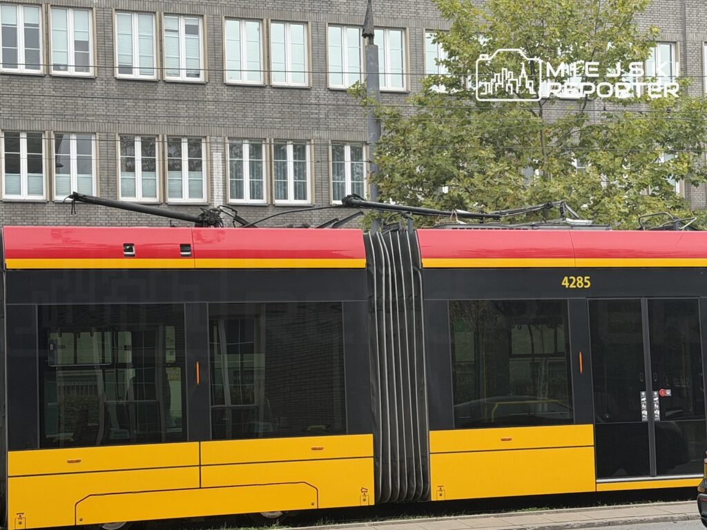 Czerwono-żółty tramwaj stojący na ulicy, z pantografem przy przewodach trakcyjnych, na tle murowanego budynku.