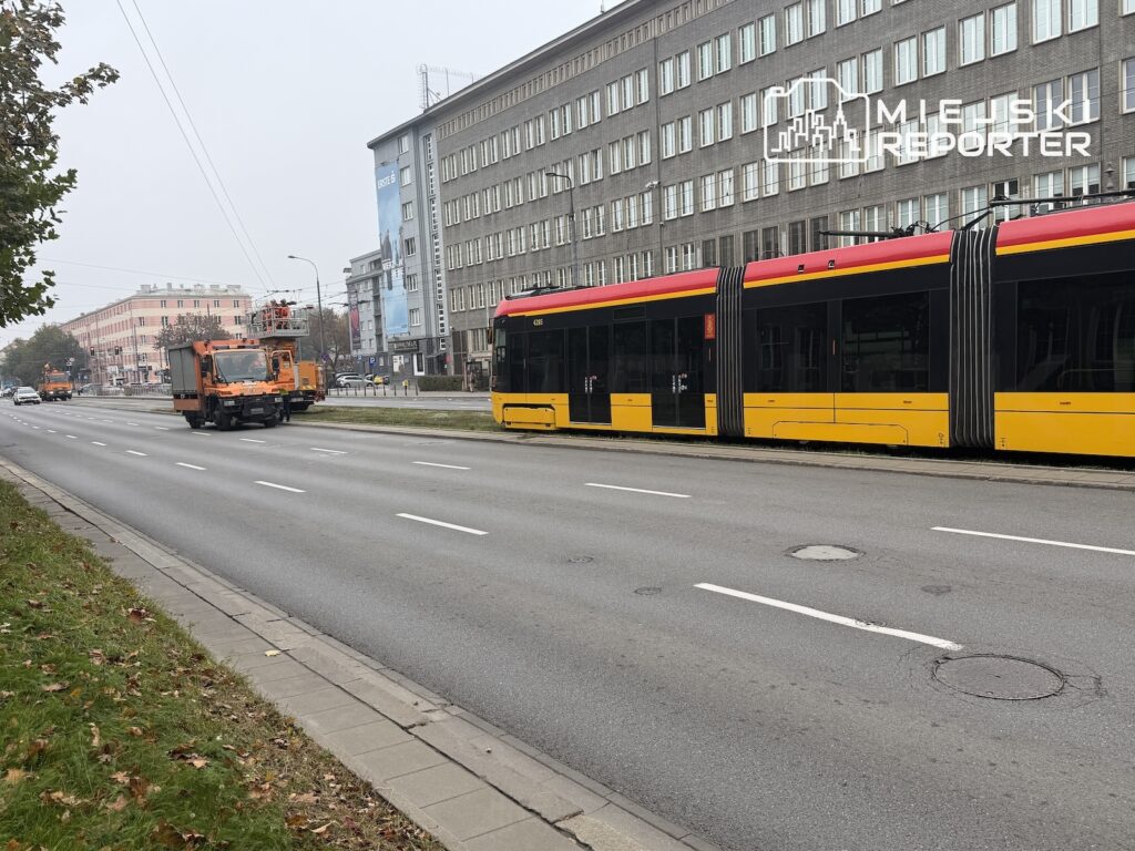 Żółto-czerwony tramwaj stoi na ulicy, a obok niego znajduje się pomarańczowy pojazd techniczny z podnośnikiem.