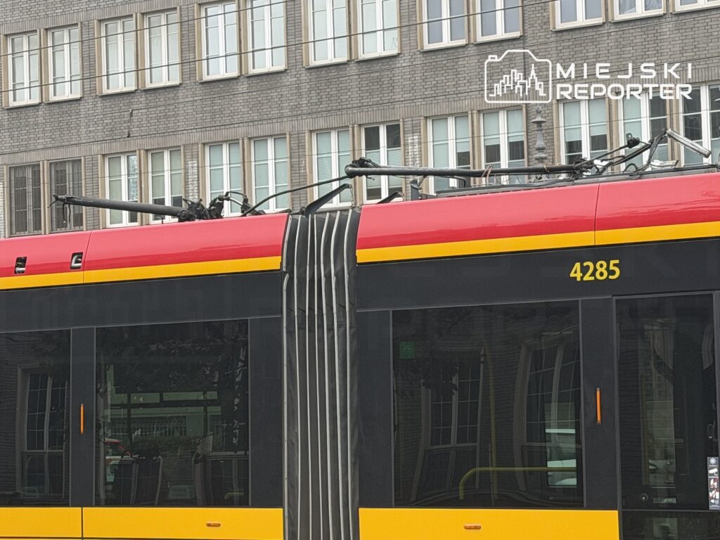 Czerwono-żółty tramwaj z uniesionym pantografem na ulicy, na tle wielopiętrowego budynku z oknami.