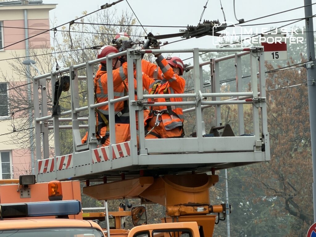 Pracownicy w pomarańczowych strojach naprawiają sieć trakcyjną z podnośnika koszowego nad ulicą.