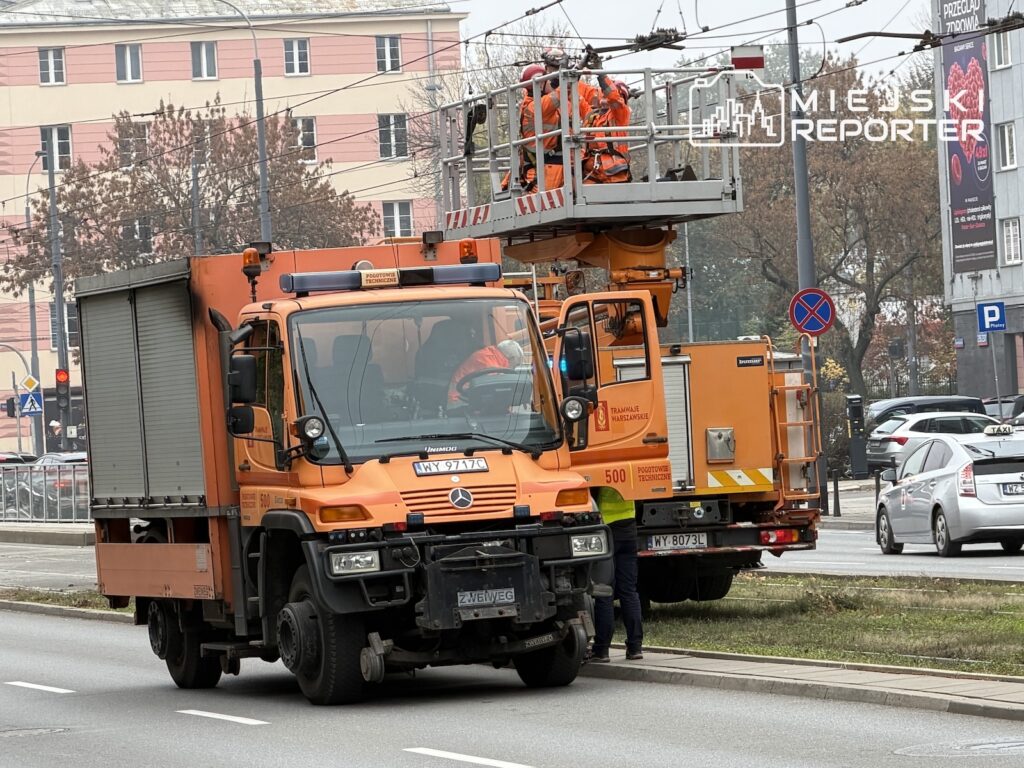 Pomarańczowe samochody techniczne i pracownicy na podnośniku przy sieci trakcyjnej nad ulicą tramwajową.