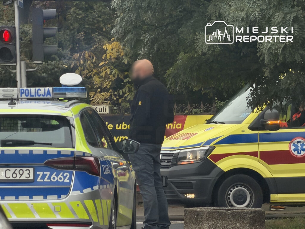 Policjant stoi między radiowozem a żółtą karetką przy skrzyżowaniu z czerwonym światłem.