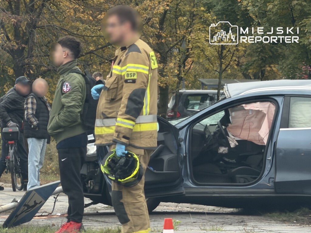 Strażak w ubraniu ochronnym stoi obok uszkodzonego samochodu z otwartymi drzwiami, przy drodze otoczonej drzewami.