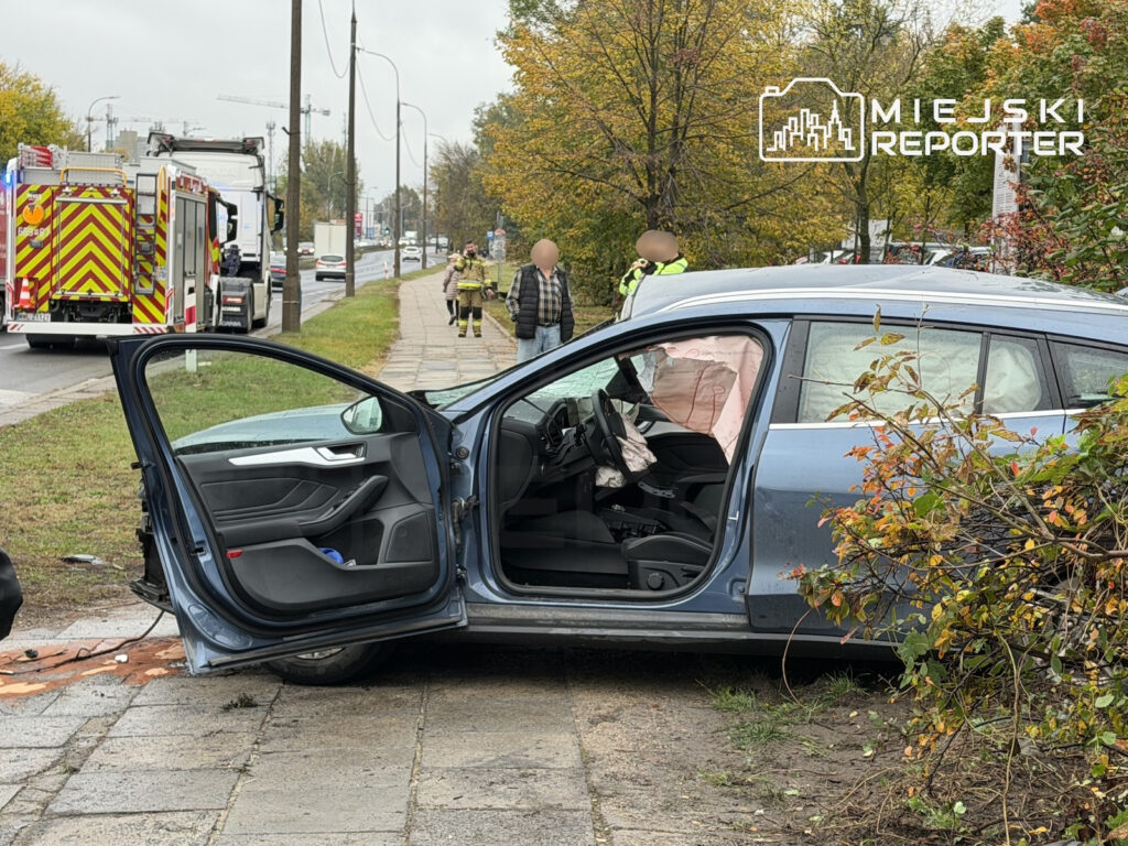 Uszkodzony samochód z otwartymi drzwiami stoi na chodniku, a obok pracują strażacy i służby ratunkowe.