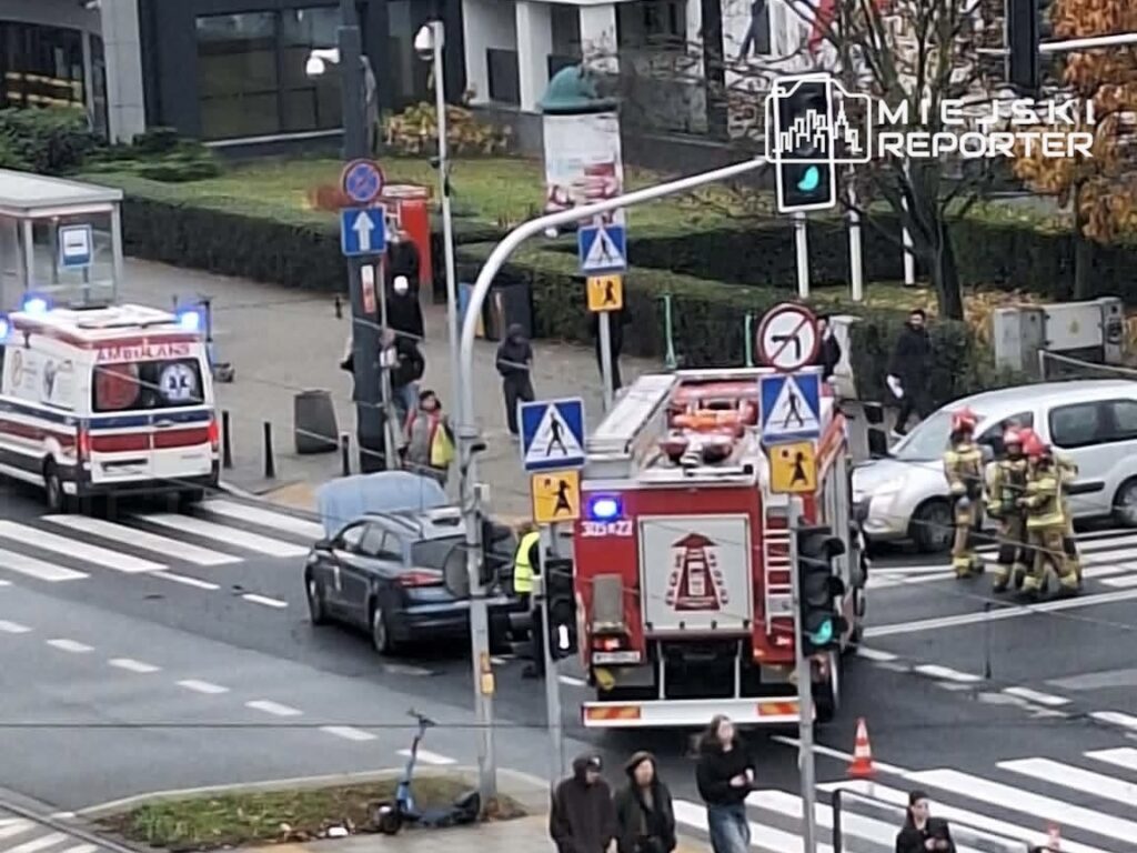 Karetka i wóz strażacki stoją na skrzyżowaniu, a strażacy pracują przy białym samochodzie osobowym.