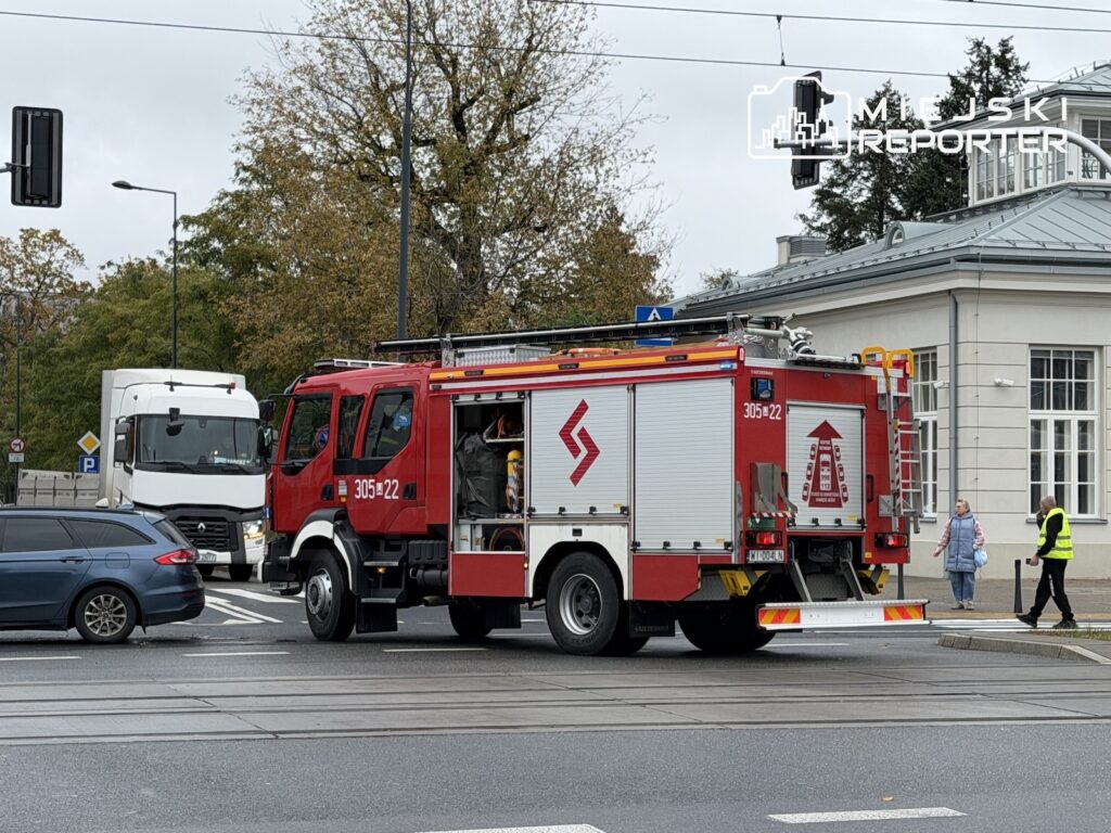 Czerwony wóz strażacki przejeżdża przez skrzyżowanie, obok samochodów i pieszych na chodniku.