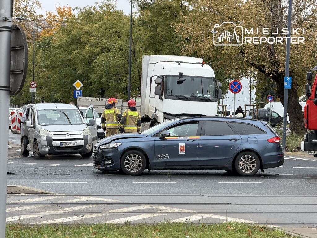 Uszkodzony granatowy samochód osobowy stoi na skrzyżowaniu obok białej ciężarówki, a strażacy zabezpieczają miejsce