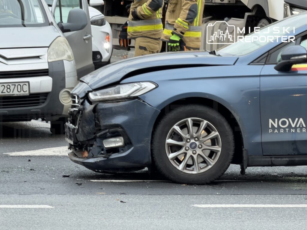 Uszkodzony przód niebieskiego samochodu osobowego po kolizji, obok stoją strażacy i zaparkowane auta.