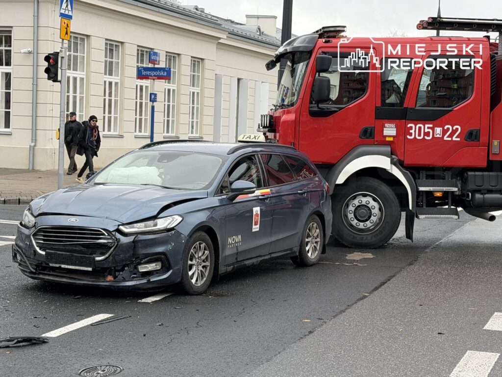 Uszkodzona taksówka stoi przed czerwonym wozem strażackim na skrzyżowaniu, obok przejścia dla pieszych.