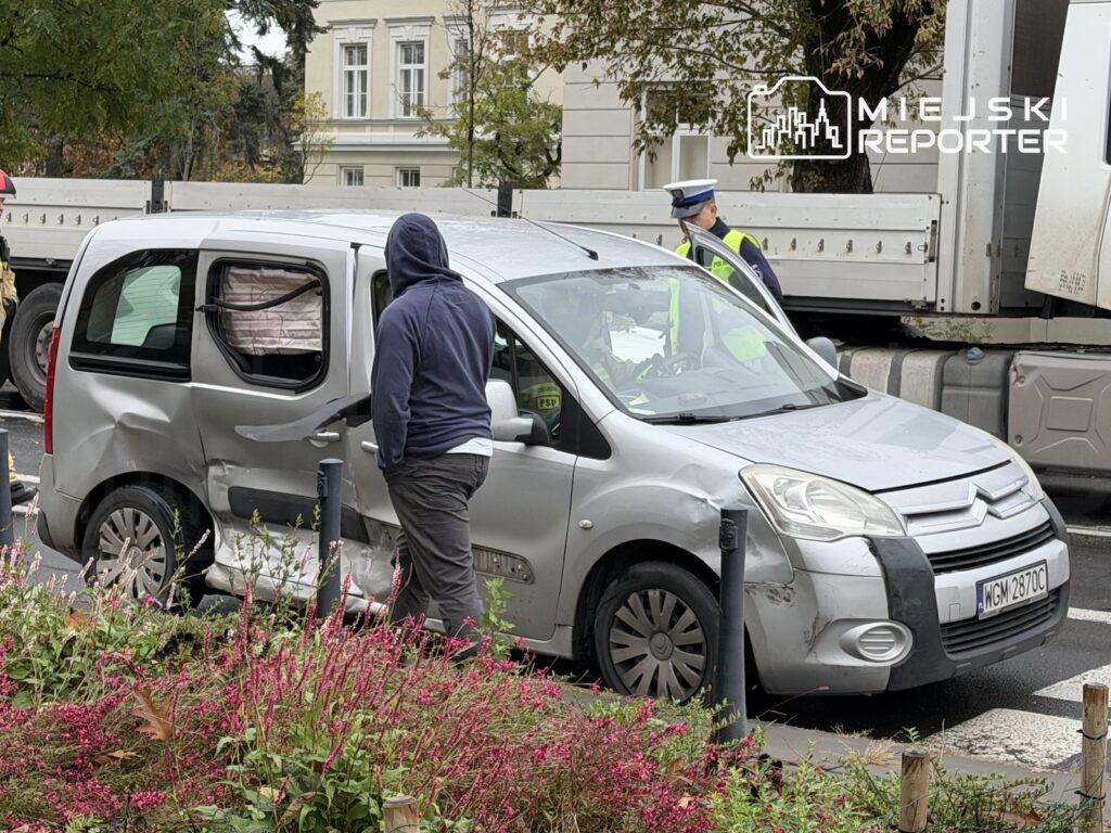 Uszkodzony srebrny samochód dostawczy stoi na ulicy obok ciężarówki, a policjant rozmawia z osobą w kapturze.