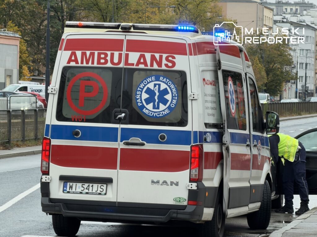 Karetka z napisem „AMBULANS” stoi na ulicy, a ratownik w kamizelce odblaskowej pochyla się przy jej boku.