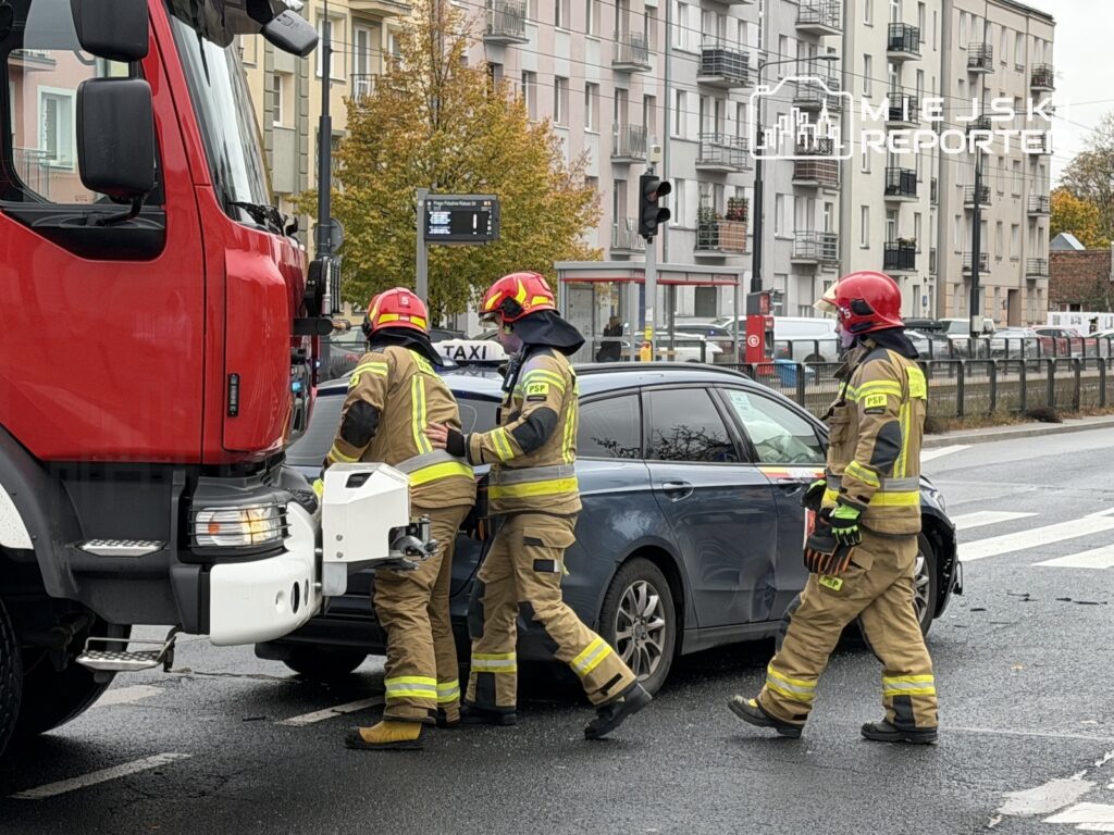 Trzech strażaków w strojach ochronnych pracuje przy czerwonym wozie i szarym samochodzie na miejskiej ulicy.