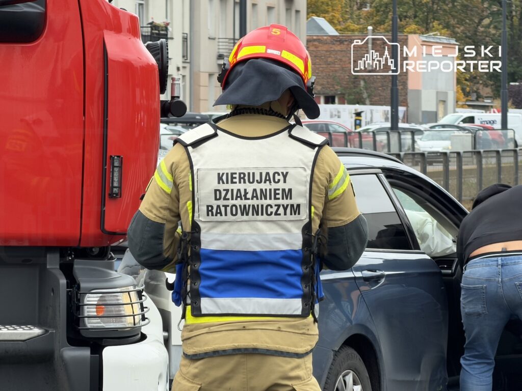 Strażak w kamizelce z napisem „KIERUJĄCY DZIAŁANIEM RATOWNICZYM” przygląda się samochodowi obok wozu strażackiego.