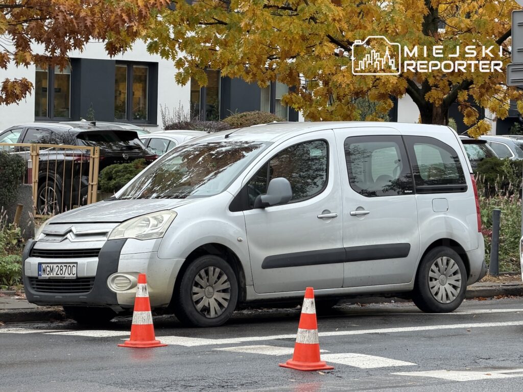 Srebrny samochód dostawczy stoi przy krawężniku, oznaczony dwoma pachołkami drogowymi pod jesiennymi drzewami.