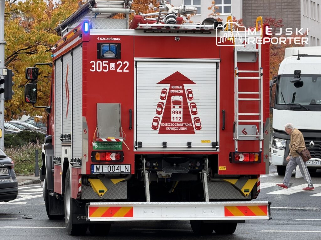 Czerwony wóz strażacki z napisem „998 112” stoi na ulicy, obok przechodzi starszy mężczyzna.