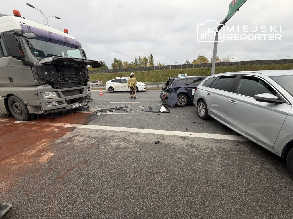Uszkodzona ciężarówka i samochody osobowe po zderzeniu na drodze, na jezdni leżą szczątki pojazdów; obok stoi ratownik.