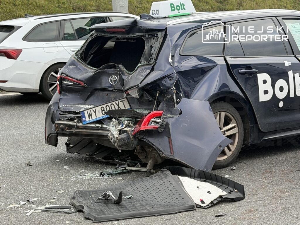 Mocno uszkodzona taksówka Bolt po zderzeniu, z rozbitą tylną szybą i elementami karoserii na jezdni.