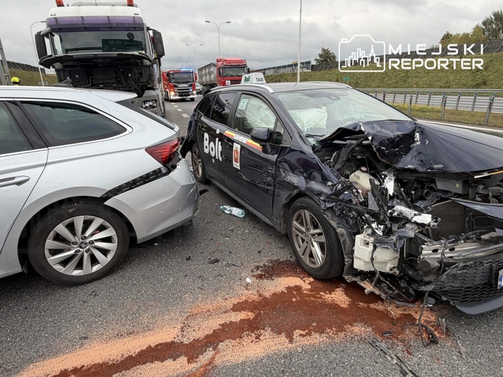 Rozbity czarny samochód taxi Bolt uderzony w tył przez srebrne auto na drodze, obok stoją ciężarówki.