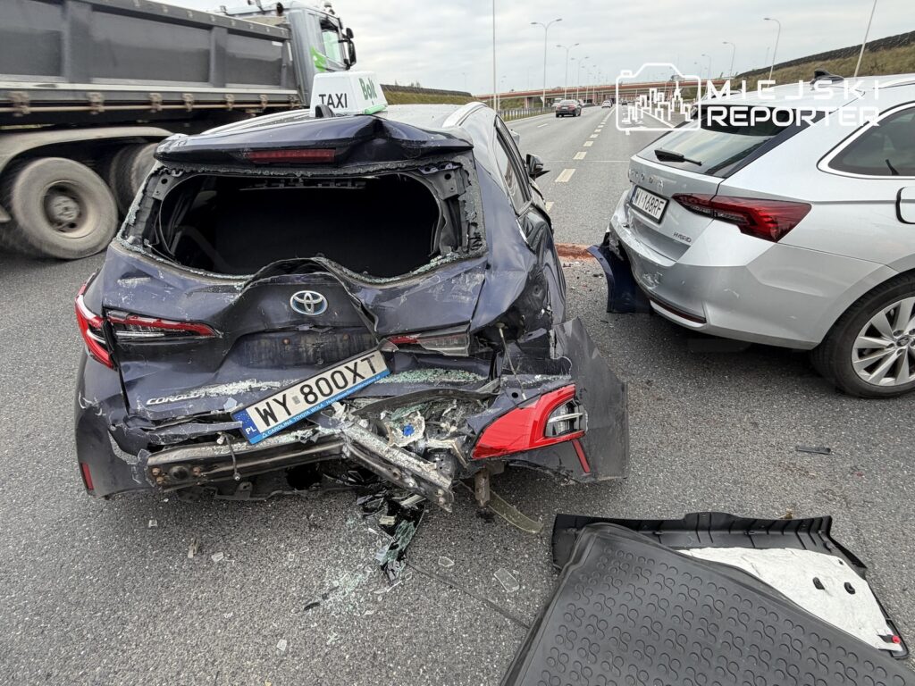 Tył mocno rozbitej Toyoty stoi na jezdni obok srebrnego auta i ciężarówki; na asfalcie leżą odłamki.