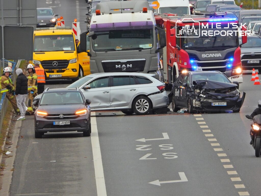 Uszkodzony czarny samochód osobowy stoi przed ciężarówką strażacką na zakorkowanej drodze; obok pracują służby ratunkowe.