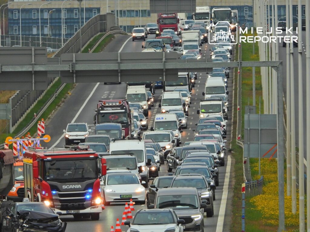 Gęsty korek na wielopasmowej drodze, wśród samochodów ciężarówka Scania i pachołki drogowe.