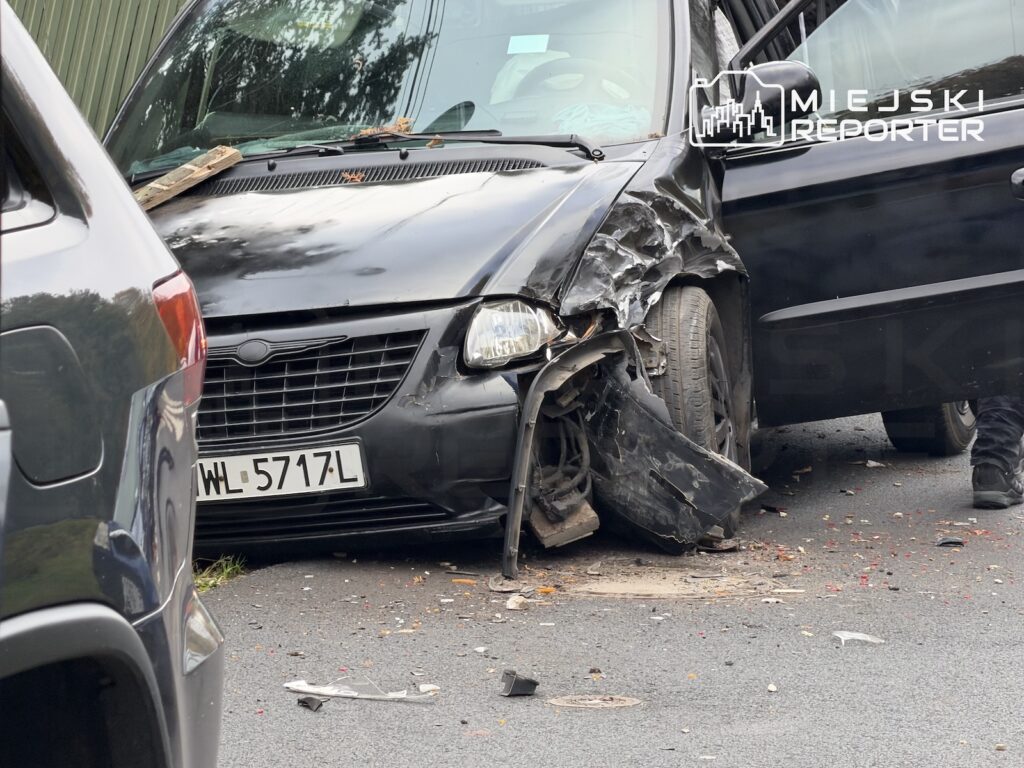 Czarny Chrysler z rozbitym przodem stoi na jezdni obok drugiego samochodu, a jego drzwi są otwarte.