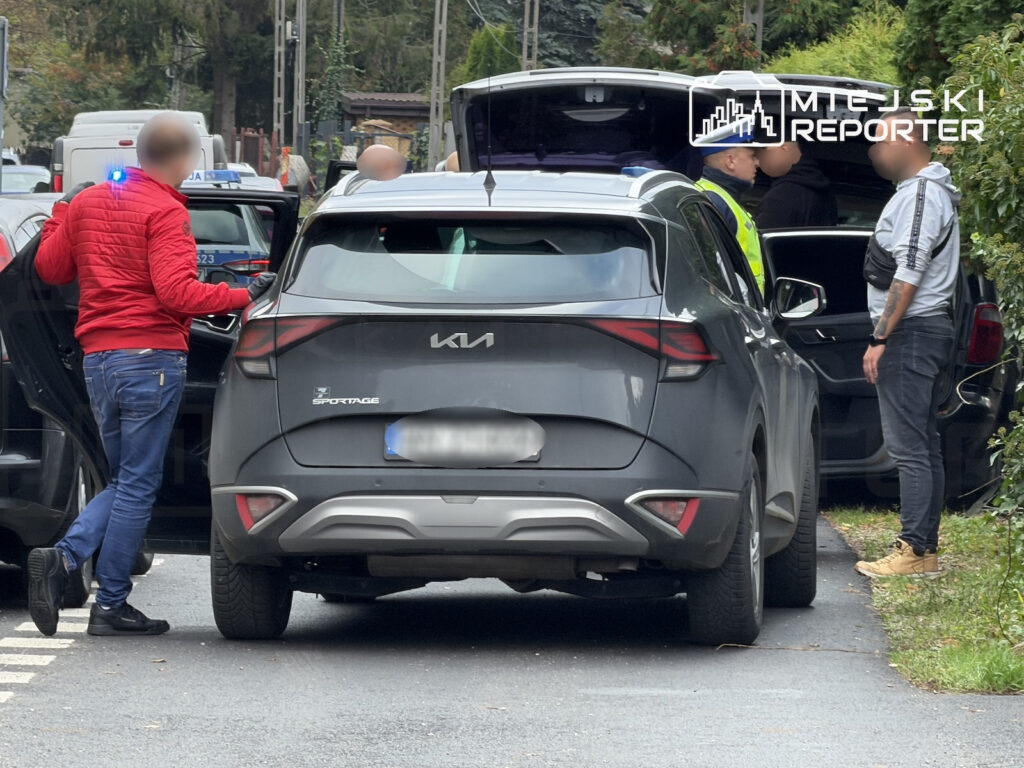 Mężczyźni stoją przy szarym SUV-ie Kia na drodze; obok zaparkowany jest policyjny radiowóz z włączonymi światłami.