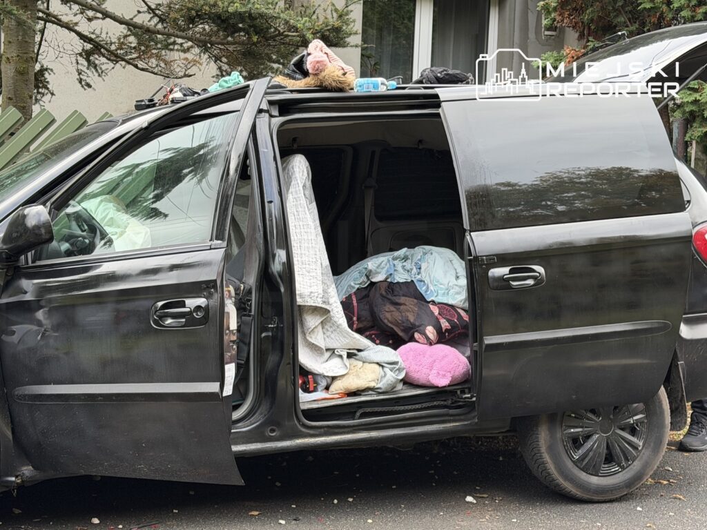 Ciemny minivan z otwartymi drzwiami, w środku i na dachu widoczne ubrania oraz pościel, zaparkowany przy budynku.