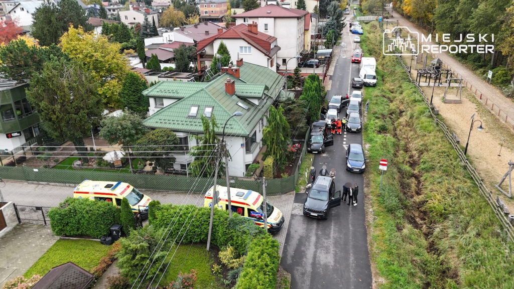Dwa ambulanse i samochody stoją na wąskiej ulicy między domami a zarośniętym torowiskiem, obok służby ratunkowe.