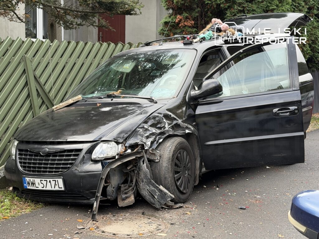 Uszkodzony czarny Chrysler z otwartymi drzwiami stoi przy zielonym ogrodzeniu posesji.