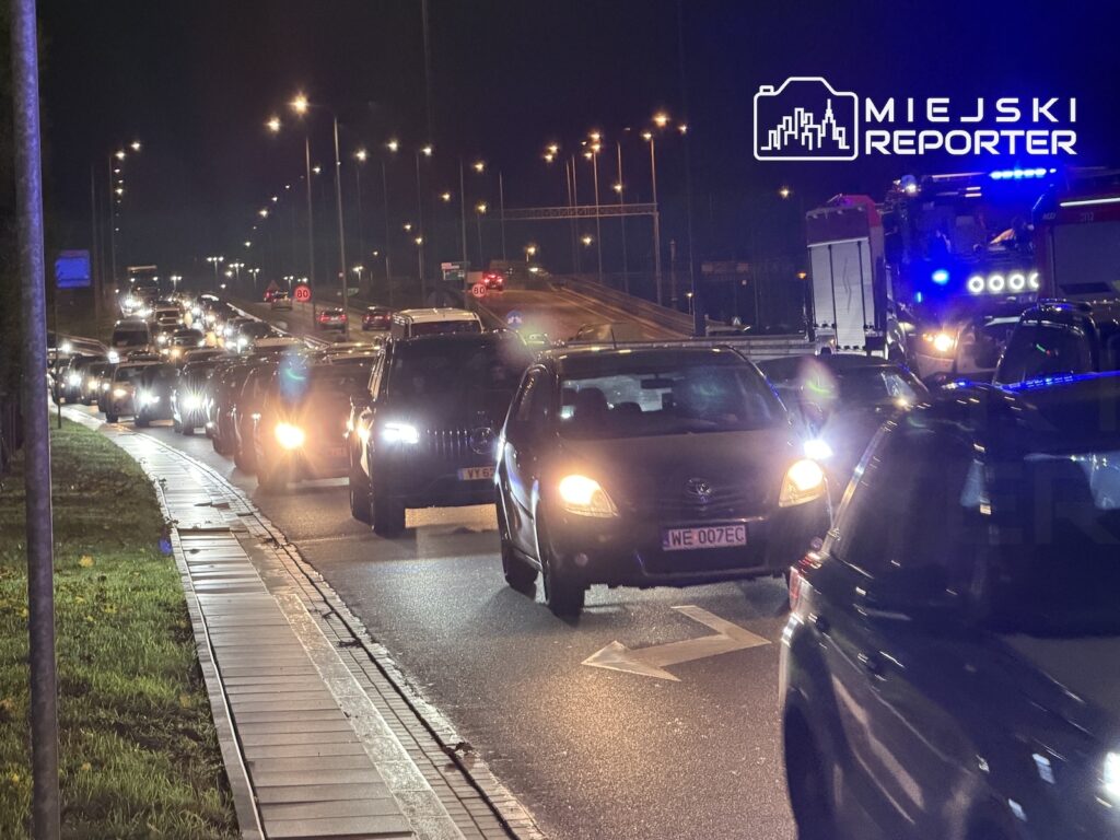 Korek samochodów na oświetlonej nocą drodze, obok pojazdów służb z włączonymi niebieskimi światłami.