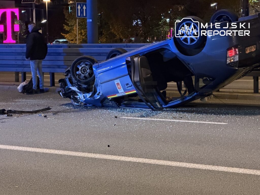 Niebieski samochód leży przewrócony na dachu na nocnej jezdni, obok stoją mężczyzna i bariera drogowa.