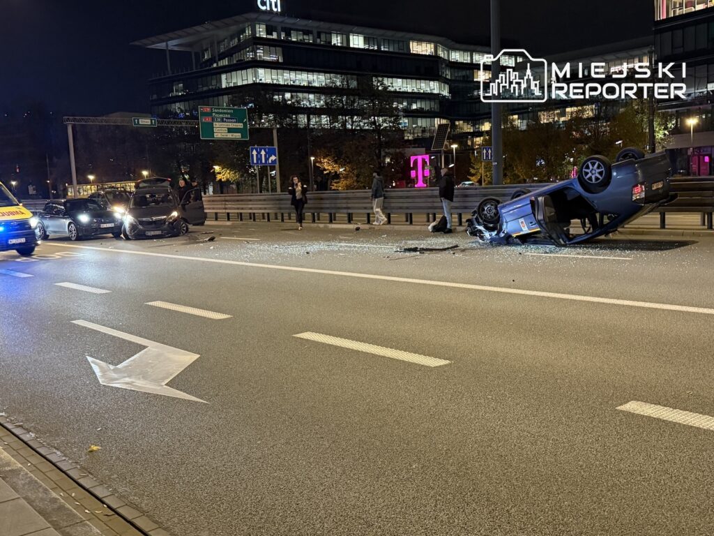Przewrócony na dach samochód stoi na nocnej ulicy; obok leżą odłamki szkła, a na jezdni są radiowozy.