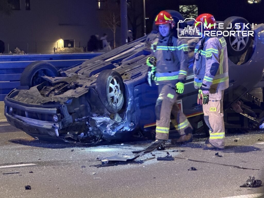 Dwóch strażaków w odblaskowych strojach stoi obok przewróconego na dach samochodu na nocnej drodze pełnej odłamków.