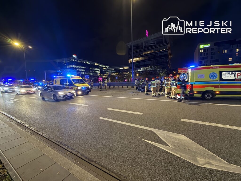 Ratownicy medyczni i strażacy stoją przy karetce na nocnej ulicy, obok pojazdów służb z włączonymi światłami.