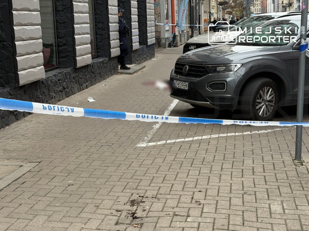 Policyjna taśma odgradza chodnik przy zaparkowanym SUV-ie; na kostce brukowej widać ciemne plamy.