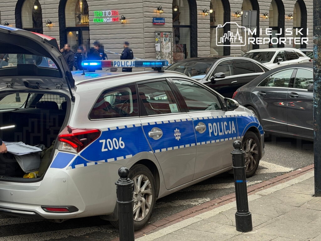 Radiowóz z napisem „POLICJA” i włączonymi światłami stoi przy ulicy, obok zaparkowanych samochodów.