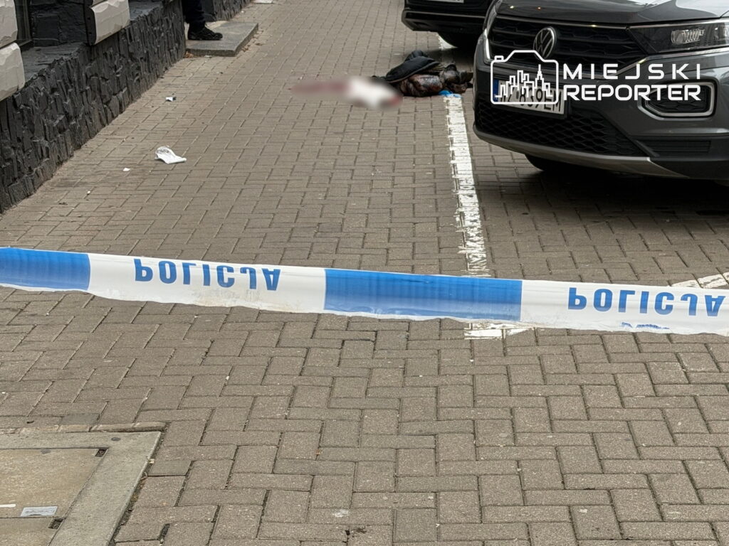 Taśma policyjna odgradza chodnik, na którym przy zaparkowanym samochodzie leży osoba.