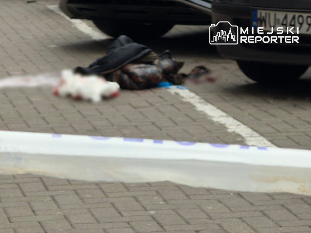 Osoba leży na chodniku obok samochodu, a jej ciało jest częściowo przykryte ciemnym materiałem; miejsce odgrodzono taśmą.
