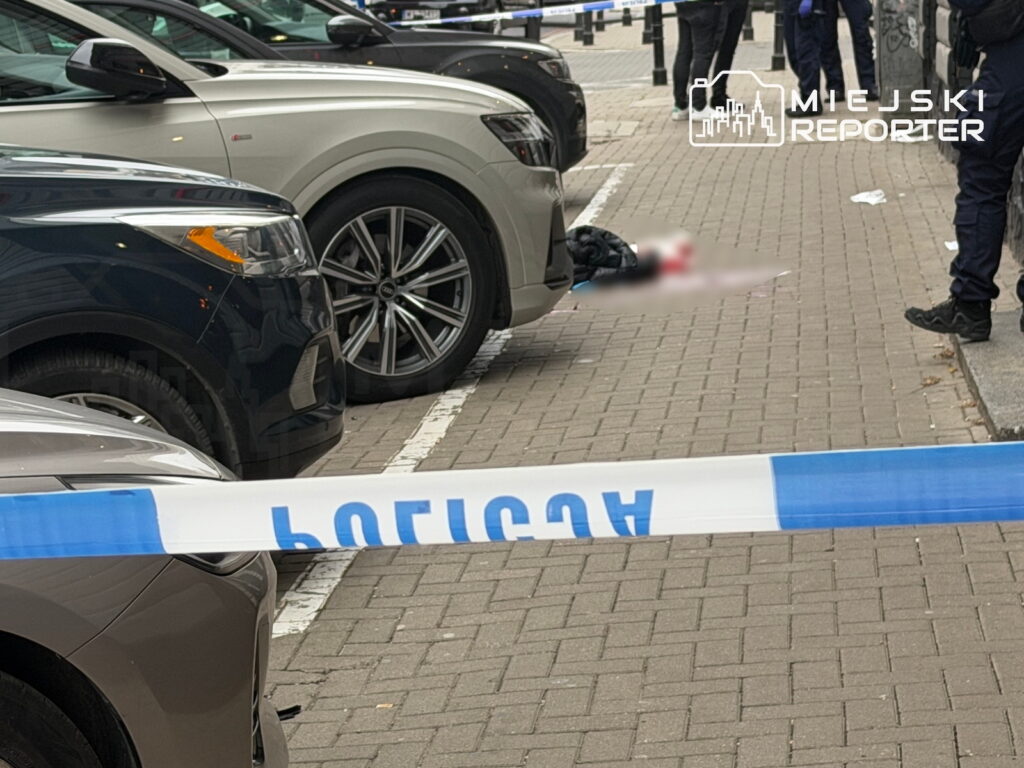 Policyjna taśma odgradza zaparkowane samochody i fragment chodnika, gdzie leży osoba zasłonięta rozmyciem.