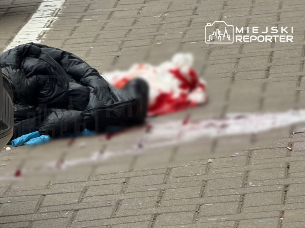 Zakrwawione ciało na chodniku obok czarnej kurtki; miejsce zdarzenia zasłonięte rozmyciem.