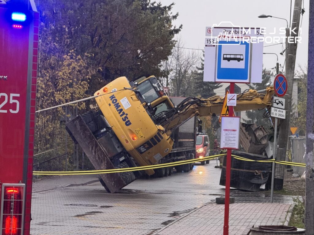 Przewrócona żółta koparka leży na boku przy przystanku autobusowym, za taśmą zabezpieczającą ulicę.