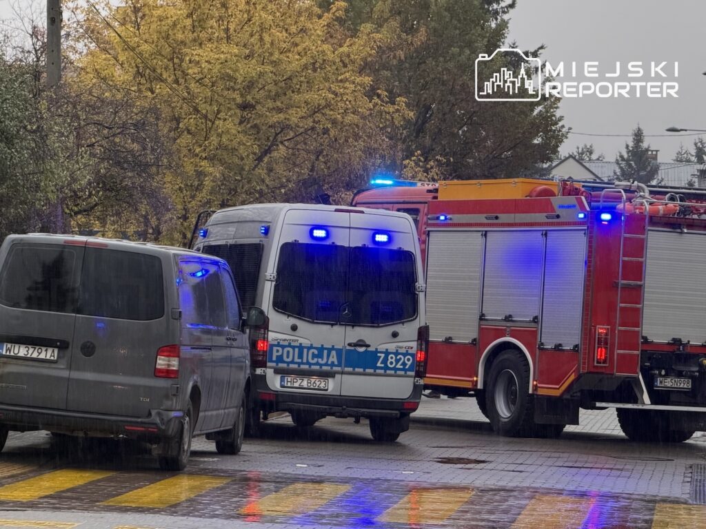 Radiowóz policyjny i czerwony wóz strażacki z włączonymi sygnałami stoją na mokrej ulicy obok zaparkowanego busa.