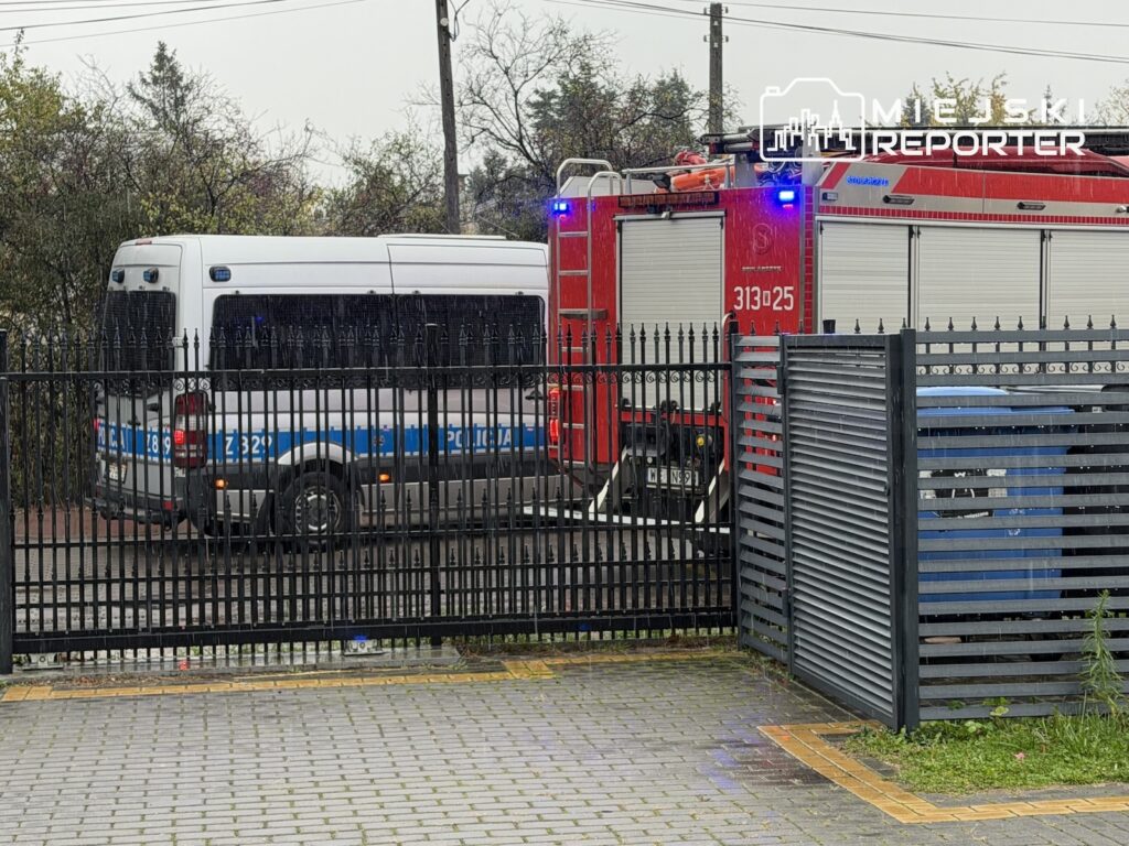Policyjny bus i czerwony wóz strażacki z włączonymi sygnałami stoją za ogrodzeniem przy ulicy.
