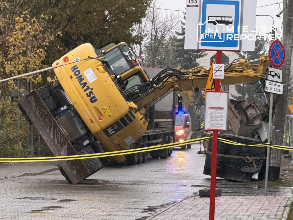 Żółta koparka Komatsu przewrócona na bok na ulicy, otoczona taśmą zabezpieczającą przy przystanku autobusowym.